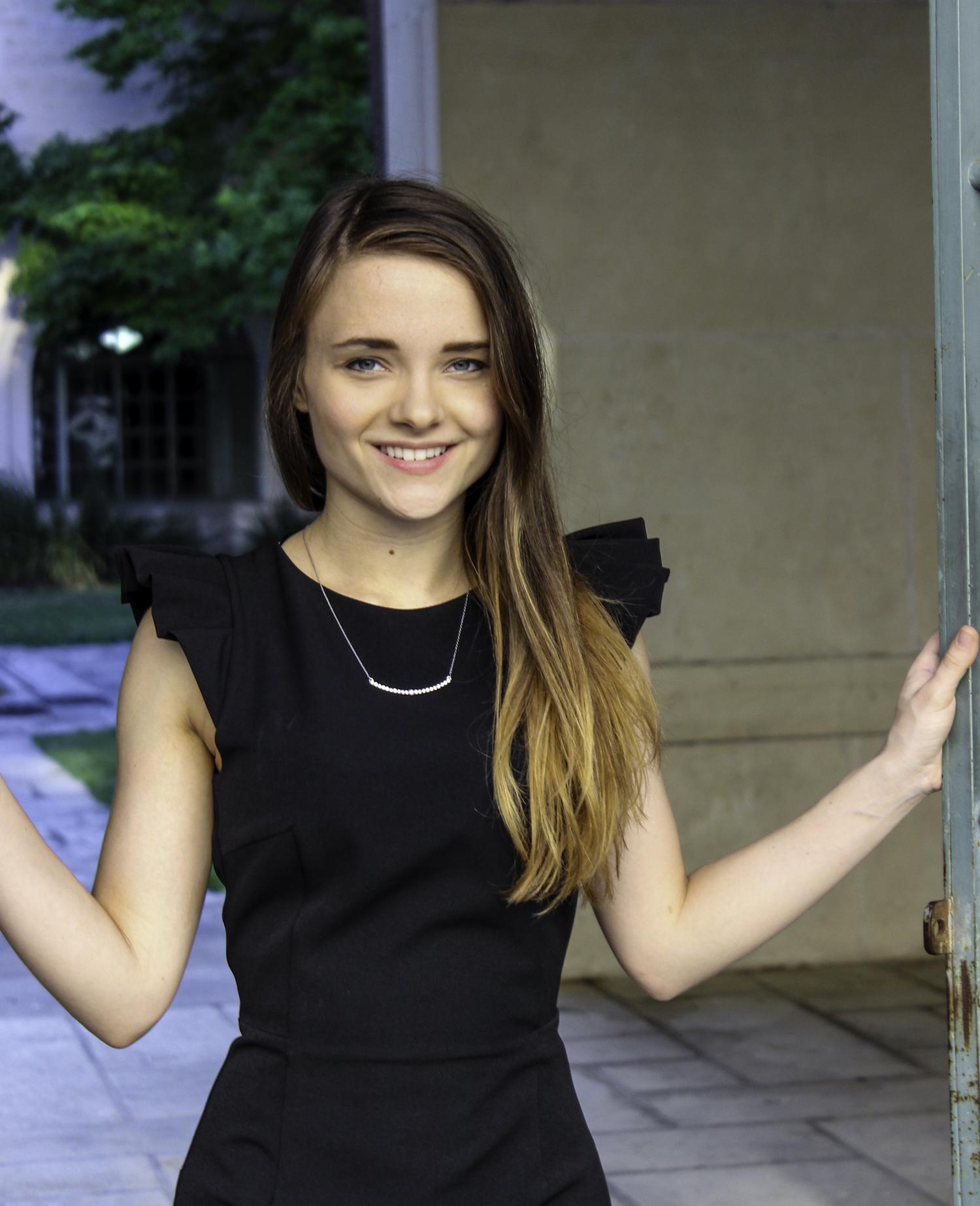 阿比盖尔纽厄尔 stands in a black dress with ruffled shoulders, 对着镜头微笑, with her hands on either side of a doorway in an outdoor space.