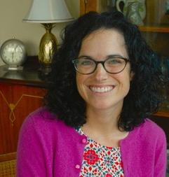 Katherine Turk in glasses, a geometric patterned white, blue and pink shirt, 还有一件亮粉色的开襟羊毛衫，背后的木边桌上放着一盏金色的灯和装饰品. 