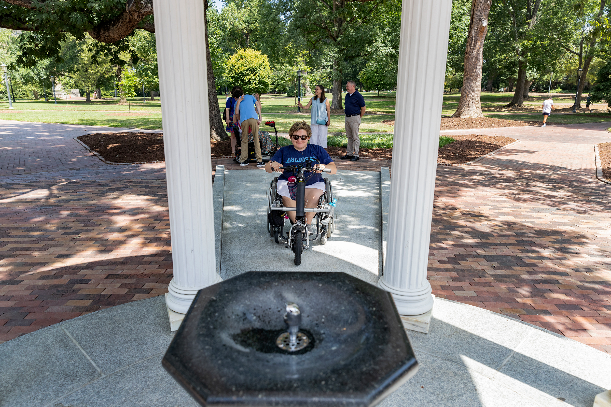 A person using a wheelchair on a sloped pathway approaches the fountain of the Old Well on the campus of UNC-Chapel Hill.