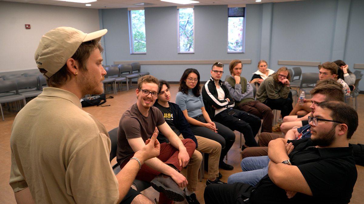 Student with white baseball cap talking in front of seated group of students.